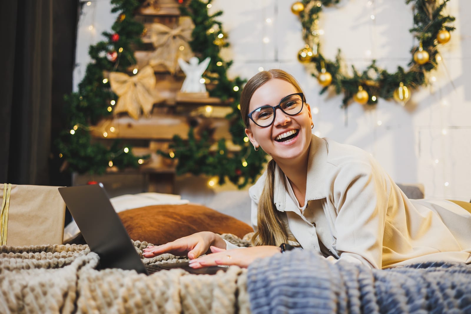 A smiling woman wearing glasses lies on a bed with a laptop in front of her. Behind her, a Christmas tree and festive wreaths decorated with lights create a cozy holiday atmosphere.