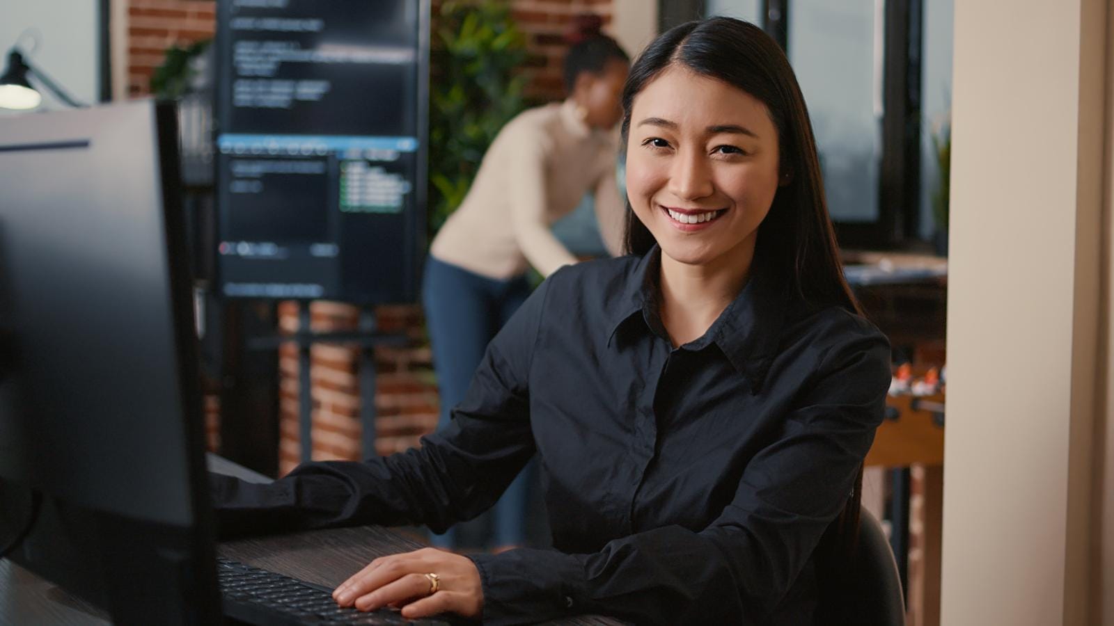 A woman in a black shirt smiles at the camera while sitting at a desk with a computer monitor; another person works in the background in a modern office setting.