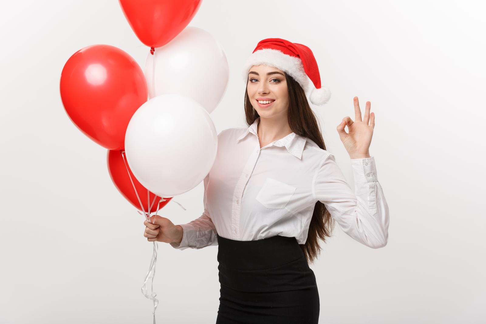 A woman wearing a Santa hat, white shirt, and black skirt holds red and white balloons in one hand and makes an "OK" gesture with the other, smiling against a plain white background.