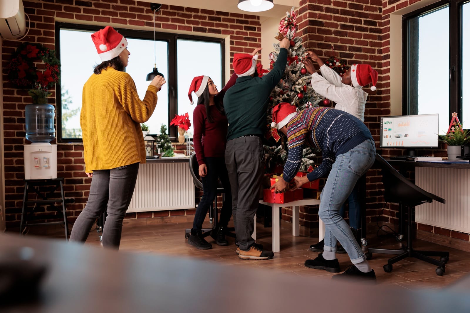 A group of people wearing Santa hats decorate a Christmas tree together in a modern office with brick walls. Gift boxes are under the tree, and a computer, chair, and water cooler are visible in the background.