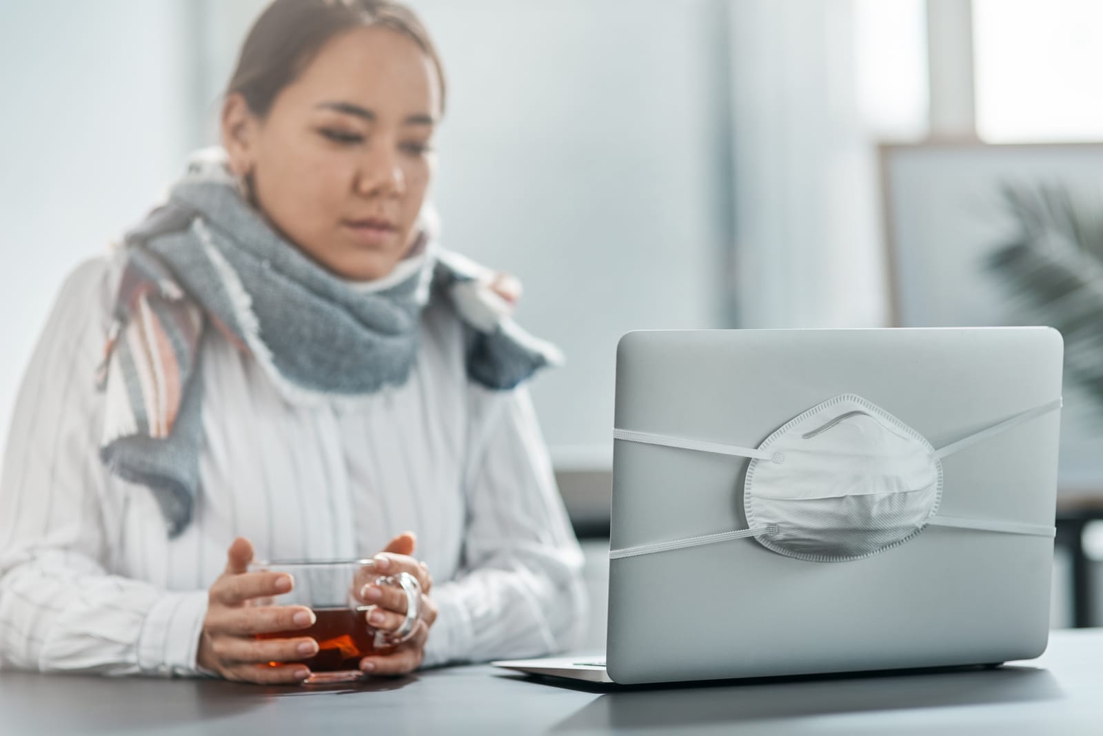 A person wearing a scarf sits at a table, holding a cup, with a laptop in front of them. A white face mask is secured over the laptop screen. The atmosphere suggests illness or a health concern.
