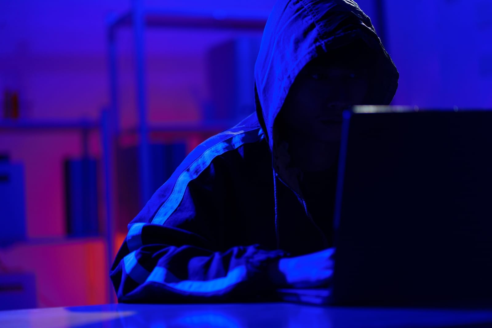 A person in a hooded jacket sits in front of a laptop in a dark room with blue and purple lighting, their face partially hidden in shadow, creating a mysterious atmosphere.